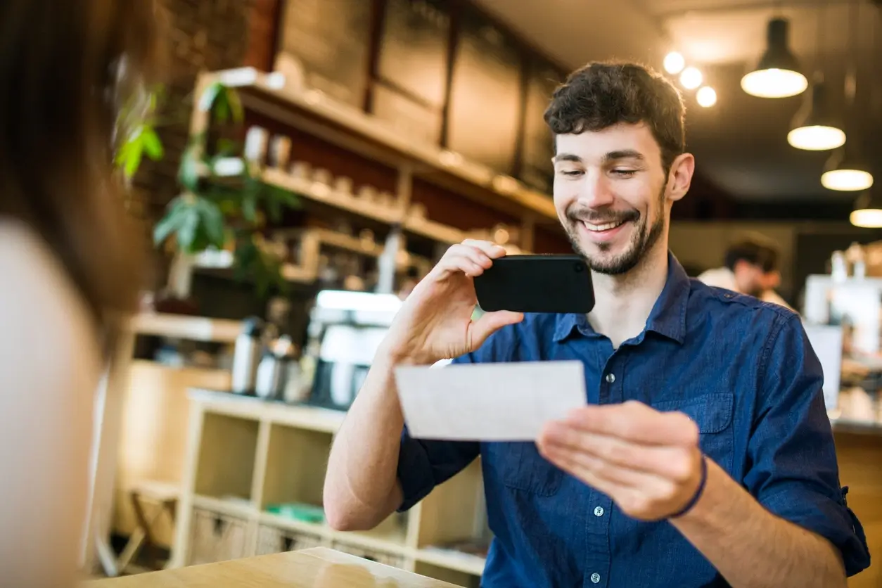 A working professional traveling at a client site smiles as he takes a photo of a receipt with his smartphone. He plans to upload the receipt using SMS and Receipt-AI, which is integrated with Xero or QuickBooks, while sitting in a cozy café environment. Indicating the ease of using the app.